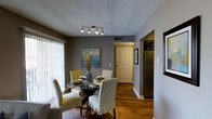 This single-floor space presents a thoughtfully designed dining area that serves as a central gathering point. The room features warm hardwood flooring that contrasts elegantly with the cool gray-toned walls, creating a balanced and inviting atmosphere. A round glass-top dining table anchors the space, surrounded by four upholstered chairs in a complementary yellow-and-white patterned fabric, adding a touch of texture and personality to the room.

Overhead, a multi-bulb track lighting fixture mounted on a textured ceiling provides focused illumination across the dining area. A large sliding glass door fitted with vertical blinds lines one wall, allowing natural light to filter into the space. A framed botanical wall art piece adds a decorative accent to the gray walls.

Adjacent to the dining area, an open passageway leads into the kitchen, where a stainless steel refrigerator is visible, hinting at a well-equipped culinary space. A framed artwork on the hallway wall adds continuity to the decor. The overall ambiance is cozy and contemporary, with a neutral color palette accented by warm wood tones and patterned textiles.