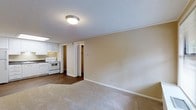 This single-floor space offers a straightforward, functional layout designed for comfortable everyday living. The main living area is finished with neutral beige carpet, providing a warm underfoot feel, while clean white baseboards frame the room and contribute to a bright, tidy aesthetic. The textured ceiling adds a classic touch overhead, and a flush-mount ceiling light provides reliable ambient illumination.

The living area flows openly into the kitchen, which transitions to durable wood-look flooring. The kitchen is equipped with white cabinetry offering ample storage, a white electric range, and an overhead range hood. A skylight above the kitchen brings in additional natural light, enhancing the overall brightness of the space.

A doorway off the main living area leads to an inner hallway, providing access to additional rooms beyond. The neutral, warm beige wall tones are consistent throughout, creating a cohesive and calm atmosphere across the space. The clean, understated design makes the space highly adaptable for a variety of furnishing styles. Overall, the layout prioritizes practicality and ease of movement, making it a well-organized and livable single-floor environment.