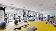 Located at Lakes at 610 in Houston, Texas, this well-equipped fitness center offers a comprehensive workout environment designed to support a wide range of exercise routines. The space is bright and welcoming, featuring large floor-to-ceiling windows along the primary wall that flood the room with natural light and provide views of the surrounding palm trees, pool area, and lush landscaping.

The flooring combines vibrant yellow carpet tiles with neutral sections, creating distinct zones throughout the space. A row of cardio machines — including treadmills, an elliptical, and a stationary bike — lines the windowed wall, allowing users to enjoy the outdoor scenery during workouts. On the opposite side, a full-wall mirror expands the sense of space and supports strength training activities.

The strength training area includes a cable machine, selectorized resistance machines for targeted muscle groups, and a dedicated free weight section with a multi-tiered dumbbell rack. Flat and adjustable benches are positioned throughout for versatile use. A wall-mounted storage rack holds medicine balls, resistance bands, and stability balls, rounding out the functional training options.

The clean white walls are accented with motivational signage, contributing to an energetic atmosphere. Recessed lighting supplements natural light to ensure the space remains well-lit throughout the day.