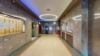 This single-floor lobby presents a well-preserved, classically designed entrance space that blends mid-century architectural character with functional building amenities. The interior is defined by richly veined marble wall cladding in warm brown and gray tones, which lines the corridors and surrounds the elevator banks, lending a sense of formality and permanence to the space.

The flooring throughout is polished terrazzo in a cream and neutral palette, bordered by a terracotta and green inlay pattern that adds visual definition along the corridors and transition zones. A striking oval ring pendant light fixture hangs centrally from the white ceiling, providing focused illumination while serving as a bold architectural centerpiece. Complementing this, ambient colored lighting—shifting between teal, blue, and purple hues—is recessed along the ceiling perimeter, creating a dynamic and atmospheric quality.

The elevator bank features multiple brass-trimmed elevator doors set into the marble walls, accompanied by a brass luggage cart, suggesting a hospitality or mixed-use function. A glazed reception or concierge window is positioned along one wall. The entrance vestibule includes full-height glass doors with metal framing, allowing views to the exterior streetscape. A stairwell with marble walls and terrazzo flooring provides additional vertical access.