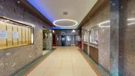 This single-floor lobby presents a well-preserved, classically designed entrance space that blends mid-century architectural character with functional building amenities. The interior is defined by richly veined marble wall cladding in warm brown and gray tones, which lines the corridors and surrounds the elevator banks, lending a sense of formality and permanence to the space.

The flooring throughout is polished terrazzo in a cream and neutral palette, bordered by a terracotta and green inlay pattern that adds visual definition along the corridors and transition zones. A striking oval ring pendant light fixture hangs centrally from the white ceiling, providing focused illumination while serving as a bold architectural centerpiece. Complementing this, ambient colored lighting—shifting between teal, blue, and purple hues—is recessed along the ceiling perimeter, creating a dynamic and atmospheric quality.

The elevator bank features multiple brass-trimmed elevator doors set into the marble walls, accompanied by a brass luggage cart, suggesting a hospitality or mixed-use function. A glazed reception or concierge window is positioned along one wall. The entrance vestibule includes full-height glass doors with metal framing, allowing views to the exterior streetscape. A stairwell with marble walls and terrazzo flooring provides additional vertical access.