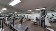 This fitness center is a dedicated exercise facility designed to support a full range of workout routines. The space is laid out as a single, open room with patterned carpet flooring that extends throughout, providing a practical surface for equipment use and light floor exercises. Overhead fluorescent lighting ensures the room is evenly and brightly illuminated throughout.

A defining architectural feature is the full-length mirrored wall that spans a large portion of the room, effectively expanding the perceived size of the space and allowing users to monitor their form during workouts. The walls are finished in a soft sage-green tone, contributing to a calm and functional ambiance.

The facility is well-stocked with cardio equipment, including multiple treadmills — among them a Vision Fitness model — elliptical trainers, and a stair climber. Strength training is supported by a multi-station cable and weight machine, along with a flat workout bench. A standalone body weight scale is also present. Informational notices and a workout schedule board are posted on the wall near the entrance, adding a practical, organized touch to the space. The room is accessed through a standard panel door, with a clearly marked exit door also visible, ensuring safe and easy navigation throughout the facility.