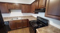 This single-floor space offers a thoughtfully designed interior centered around a functional and visually appealing kitchen. The kitchen is a standout feature, showcasing rich, dark-stained wood cabinetry with raised-panel detailing that extends to both the upper and lower sections, providing generous storage throughout. The countertops feature a warm granite-style finish that complements the cabinetry tones, creating a cohesive and classic aesthetic. A double-basin stainless steel sink is neatly integrated into the layout, positioned conveniently along the perimeter. The kitchen is equipped with a black electric range with coil burners and a matching overhead range hood, offering a clean, coordinated appliance package. The cabinetry wraps around the kitchen in an L-shaped configuration, maximizing workspace and storage efficiency. Underfoot, warm wood-tone flooring adds to the inviting atmosphere and ties the space together. The walls are finished in a clean, neutral tone that brightens the interior and provides a crisp contrast to the darker cabinetry. Overall, the space presents a well-balanced combination of practicality and style, making it a comfortable and efficient environment for everyday living.