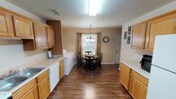 This two-floor residence offers a practical and comfortable layout with warm, neutral tones and durable hardwood-style flooring carried throughout the main living areas. The galley-style kitchen is well-equipped, featuring honey-toned wood cabinetry, light countertops, white appliances including a dishwasher and range, and ample upper and lower cabinet storage. The kitchen flows seamlessly into a combined dining and laundry area, where a window dressed with grommet-top curtains brings in natural light. A pendant chandelier highlights the dining space, which comfortably accommodates a round table and chairs. Side-by-side washer and dryer units are conveniently integrated into this area. The living room is an inviting, open space with a ceiling fan, a front entry door with sidelights, and windows providing natural light. Carpet-floored bedrooms offer a quiet retreat, each with windows, neutral walls, and ceiling-mounted light fixtures. The upper-floor hallway features carpeted flooring and thoughtful decorative touches. Bathrooms are finished with wood-toned vanity cabinetry, white fixtures, and hardwood-style flooring, with one bathroom offering a full tub-shower combination. Throughout the home, a consistent, understated aesthetic creates a cohesive and functional living environment.