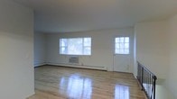 This two-floor space offers a straightforward, functional layout with classic design elements that create a bright and welcoming environment. The main living area is defined by gleaming hardwood floors that reflect natural light, contributing to an open and airy atmosphere. Large windows along the exterior wall allow generous amounts of daylight to fill the room, providing views of the surrounding greenery outside. A charming panel door with a grid-style window adds a traditional architectural touch and serves as an additional source of natural light. The walls are finished in clean, neutral white tones, lending a versatile backdrop suitable for a variety of uses and furnishing styles. A baseboard heating system runs along the perimeter of the main room, accompanied by a window-mounted air conditioning unit, ensuring year-round climate comfort. The overall aesthetic is simple and unfussy, emphasizing functionality and livability. With its polished floors, abundant natural light, and classic details, this two-floor space presents a solid foundation that is both practical and adaptable to a range of needs.