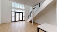 This thoughtfully designed two-story space showcases a striking contemporary aesthetic with clean lines and an open, airy atmosphere. The main level immediately impresses with its dramatic double-height ceilings and an expansive wall of floor-to-ceiling windows fitted with roller shades, flooding the interior with abundant natural light. Light-toned hardwood flooring runs throughout the main living areas, complementing the crisp white walls and creating a warm, inviting atmosphere.

A modern staircase with sleek black cable railings connects the two levels, adding an industrial-chic design element that is both functional and visually striking. The kitchen is well-appointed with dark slate-toned cabinetry, open wood shelving, a white quartz countertop island, subway tile backsplash, and a full suite of stainless steel appliances, offering both style and practicality.

The upper level features plush carpeted rooms, including a spacious bedroom with a closet, and an open loft area overlooking the main floor through matching cable railings. The bathroom is finished with dual undermount sinks set in a white quartz vanity, grey cabinetry, brushed gold hardware, large-format tile flooring, and a soaking tub. Throughout, the space balances modern design with functional living across both floors.