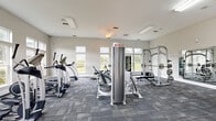 Located at Baldwin Park Drive in Tallahassee, Florida, this fitness center offers a well-appointed workout facility designed to support a full range of exercise routines. The single-floor space is thoughtfully laid out to accommodate a diverse selection of equipment without feeling cluttered.

Large, multi-pane windows line multiple walls, flooding the interior with natural light and offering pleasant views of the surrounding greenery. The bright, neutral-toned walls and patterned carpet tile flooring contribute to a clean, modern aesthetic throughout. Ceiling fans provide additional air circulation, enhancing comfort during workouts.

The cardio area is stocked with treadmills, elliptical machines, and a recumbent bike, offering a variety of low- and high-impact options. A dedicated strength training zone features a power rack with a barbell and weight plates, a multi-function cable machine, and a leg press/calf raise station. Free weights, including a full rack of dumbbells and a set of medicine balls, are also available for functional training. Wall-mounted mirrors expand the sense of space and allow for proper form checks during exercise.

A set of double glass doors provides convenient access, while posted fitness center rules and a wall-mounted TV add functional touches to the space. The overall environment is bright, organized, and purpose-built for effective fitness use.