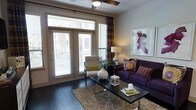 This well-appointed single-floor space combines modern design with comfortable living. The living area serves as the centerpiece, showcasing rich dark hardwood floors that contrast beautifully with neutral-toned walls, creating a warm and inviting ambiance. A bold deep-purple sofa anchors the seating area, complemented by a patterned accent chair and a sleek coffee table, reflecting a thoughtfully curated interior aesthetic.

Large French doors flood the space with abundant natural light, offering views of the exterior and providing a seamless connection to the outdoors. The doors are framed by elegant patterned drapes that add texture and visual interest to the room. Horizontal blinds on all windows allow for flexible light control throughout the day.

The ceiling features a contemporary ceiling fan, adding both functionality and a modern touch to the overall design. A patterned area rug defines the living space, layering warmth over the hardwood flooring beneath. Wall art and decorative accessories, including a sculptural vase and curated books on the coffee table, contribute to the polished, magazine-ready feel of the interior. The single-floor layout offers an efficient and cohesive flow, making it an accessible and practical living environment.