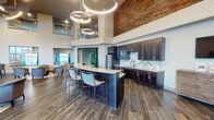 This single-floor amenity space is defined by its dramatic soaring ceilings and open, multi-functional layout. The central kitchen and lounge area serves as the focal point, showcasing dark espresso cabinetry, granite-topped counters, and a chevron-patterned tile backsplash. A large island with bar-height seating encourages social gathering, while stacked circular pendant lights add a striking architectural element overhead. A floor-to-ceiling stacked stone accent wall introduces a bold, textural contrast against the crisp white walls. Wood-look tile flooring flows continuously throughout, lending warmth and cohesion across zones. The billiards area is bathed in natural light from an expansive floor-to-ceiling glass window wall, offering outdoor views. Adjacent lounge seating areas feature curated furnishings and artwork, creating intimate conversation nooks. Interior glass window walls provide visual connectivity to a fully equipped fitness center and a dedicated co-working space fitted with workstations and modern lighting. A high-top bistro table with upholstered seating offers an additional casual work or dining option. Together, these distinct yet interconnected zones create a sophisticated, amenity-rich environment designed for both productivity and leisure.