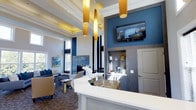 This well-appointed single-floor space makes a strong first impression through its thoughtful design and refined aesthetic. The interior is defined by dramatically high ceilings with decorative white beam detailing and a tray ceiling element that adds architectural depth. A bold navy blue accent wall contrasts elegantly against the soft neutral gray tones throughout, creating a sophisticated and cohesive color palette.

A central reception counter finished in crisp white with a granite surface serves as the focal point of the main area. Behind it, a built-in shelving unit with wood-toned paneling provides functional display space, while a wall-mounted flat-screen television adds a modern touch. Striking gold cylindrical pendant lights hang from the ceiling, providing warm, ambient illumination that complements the overall design.

Adjacent to the reception area, a comfortable lounge space features plush seating in coordinating navy and neutral tones, arranged around a fireplace. Large windows allow natural light to filter in, enhancing the open, airy feel of the space. Abstract artwork on the walls and carefully selected décor details contribute to the polished, welcoming ambiance throughout.