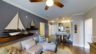 Located at 1015 Nutt St in Wilmington, NC, this single-floor residence offers a thoughtfully designed open-concept layout that seamlessly connects the living, dining, and kitchen areas. The living room features warm hardwood-style flooring, crown molding, and a ceiling fan with wood-tone blades, creating a comfortable and polished atmosphere. Coastal-inspired accents, including a decorative sailboat wall piece and floating wood shelf, lend a charming nautical character to the space. The dining area accommodates a rustic wood table surrounded by modern chairs, positioned conveniently adjacent to the kitchen. The kitchen is well-equipped with stainless steel appliances, including a refrigerator and microwave, complemented by light cabinetry, a stylish tile backsplash, and pendant lighting over the counter. Track lighting overhead ensures the cooking and dining areas are well-illuminated. The color palette features cool grey accent walls paired with white trim and ceilings, contributing to a clean and contemporary aesthetic throughout. The open sightlines across the living and kitchen spaces enhance the sense of spaciousness, making this single-floor layout both functional and visually inviting.