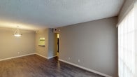This single-story, one-bedroom, one-bathroom unit offers a well-organized layout with distinct living, dining, kitchen, and sleeping areas. The open living and dining space features dark hardwood-style flooring throughout, neutral gray walls, and a textured ceiling. A pendant light fixture illuminates the dining area, while a large window fitted with vertical blinds allows natural light to fill the room. A pass-through counter connects the dining area to the kitchen, enhancing the open feel of the space.

The kitchen is equipped with white shaker-style cabinetry, granite-patterned countertops, a stainless steel sink, and black appliances including a refrigerator, dishwasher, and range. Matching hardwood-style flooring continues into the kitchen, and a flush-mount ceiling light provides bright, even illumination.

The bedroom is carpeted in a neutral tone, offering a comfortable contrast to the harder surfaces in the main living areas. A ceiling fan with light kit and vertical blinds on the window complete the room.

The bathroom features a full-size soaking tub with a white tiled surround and shower, a toilet, and granite-style countertops, maintaining a clean, cohesive aesthetic throughout the unit.