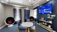 Located at 221 Fairforest Way in Greenville, SC, this thoughtfully designed community space serves as both a leasing lounge and collaborative workspace. The single-room layout makes efficient use of its compact footprint, delivering a polished, multi-functional environment with a contemporary aesthetic.

The space is anchored by a striking navy blue accent wall adorned with a bold geometric pattern, providing a dramatic backdrop for a wall-mounted display screen and a sleek console table styled with fresh orchids and decorative accents. Opposite, a business-themed wall graphic adds a creative, tech-forward energy to the room.

At the center, a marble-topped round table is surrounded by navy blue and gold rolling chairs, creating an inviting spot for meetings or consultations. Distinctive pod-style seating chairs with orange accent pillows are positioned throughout, offering private nooks for focused work or conversation. Navy drapes frame the windows, which allow natural light to filter into the space.

A standout feature is the illuminated Eco Boutique Market kiosk near the entrance, stocked with beverages and snacks for added convenience. The multicolored patterned carpet, recessed lighting, and tray ceiling details complete the space, contributing to an atmosphere that is both professional and welcoming.