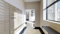 This single-floor lobby space presents a clean, contemporary design characterized by neutral beige walls and high ceilings that contribute to an open, airy atmosphere. Abundant natural light streams through large windows, casting bright reflections across the polished white and dark-bordered tile flooring, enhancing the sense of spaciousness throughout the area.

A prominent feature of the lobby is the built-in wall of numbered residential mailboxes finished in a brushed metallic tone, providing organized mail access for building occupants. The units are neatly arranged in rows, offering a practical and visually streamlined solution integrated directly into the wall.

The corridor extends beyond the mailbox area through an open archway, leading to additional common spaces within the building. This passage is well-lit, with recessed ceiling lighting complementing the natural light that filters in from the windows. A dark baseboard trim lines the lower walls, providing a sharp contrast to the lighter wall tones and adding a polished, finished look to the overall design. The space feels functional yet welcoming, with a modern aesthetic consistent throughout.