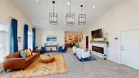 Located in Millsboro, Delaware, this single-floor community amenity space offers two distinct areas designed for relaxation and fitness. The living room is a standout, boasting soaring vaulted ceilings adorned with three dramatic black cage-style pendant chandeliers that anchor the open, airy layout. A sleek electric fireplace with a rustic wood mantle serves as the room's focal point, topped by a wall-mounted television. Seating areas are thoughtfully arranged with a mix of leather and upholstered sofas, accent chairs, and patterned area rugs, creating an inviting and stylish atmosphere. Large windows dressed in rich navy drapes flood the space with natural light, while an arched pass-through connects to a small bar or lounge area in the background. The exercise room features bold terracotta-toned walls accented with motivational typography, a distinctive windmill-style ceiling fan, and skylights that brighten the space. It is well-equipped with treadmills, ellipticals, a cable machine, stationary bikes, a weight bench, and an organized rack of exercise balls and accessories, offering a comprehensive fitness experience within a vibrant, energizing environment.