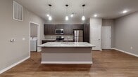 This well-appointed single-floor unit offers a thoughtfully designed open-concept layout that seamlessly connects the main living and kitchen areas. The kitchen serves as the centerpiece of the space, showcasing a large granite-topped island with an integrated sink, dark espresso cabinetry, and a full suite of stainless steel appliances including a refrigerator, range, and microwave. Pendant lighting above the island adds a modern touch, while recessed ceiling lights illuminate the space throughout. Warm hardwood-style flooring runs through the kitchen and living areas, lending a cohesive and contemporary feel. The living area is generously sized with direct access to an outdoor space via a glass-paneled door, along with a window that brings in natural light. The bedrooms are finished with plush wall-to-wall carpet and fitted with roller shade window treatments, with one bedroom featuring dual windows and a ceiling fan. The bathroom includes a dark-stained vanity with a white countertop, a well-lit mirror, and a walk-in shower with chrome fixtures. An adjacent walk-in closet provides ample storage. A dedicated laundry area houses a full-size top-loading washer and dryer, adding everyday convenience to this move-in-ready space.