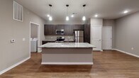 This well-appointed single-floor unit offers a thoughtfully designed open-concept layout that seamlessly connects the main living and kitchen areas. The kitchen serves as the centerpiece of the space, showcasing a large granite-topped island with an integrated sink, dark espresso cabinetry, and a full suite of stainless steel appliances including a refrigerator, range, and microwave. Pendant lighting above the island adds a modern touch, while recessed ceiling lights illuminate the space throughout. Warm hardwood-style flooring runs through the kitchen and living areas, lending a cohesive and contemporary feel. The living area is generously sized with direct access to an outdoor space via a glass-paneled door, along with a window that brings in natural light. The bedrooms are finished with plush wall-to-wall carpet and fitted with roller shade window treatments, with one bedroom featuring dual windows and a ceiling fan. The bathroom includes a dark-stained vanity with a white countertop, a well-lit mirror, and a walk-in shower with chrome fixtures. An adjacent walk-in closet provides ample storage. A dedicated laundry area houses a full-size top-loading washer and dryer, adding everyday convenience to this move-in-ready space.