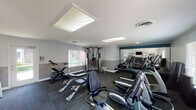 This fitness center is a thoughtfully designed, single-floor space offering a comprehensive range of workout equipment to support a variety of exercise routines. The room features neutral two-tone walls with a gray lower half and light upper half, accented by a teal stripe that adds a modern touch. Crown molding and flush lighting panels contribute to a clean, polished aesthetic throughout.

The cardio section is well-stocked with True-brand treadmills, stationary recumbent and upright bikes, and elliptical machines. A multi-functional cable machine provides options for a wide range of strength exercises, while a dedicated free weights area houses a full rack of dumbbells alongside adjustable workout benches. Stability balls and exercise mats are also available for core and flexibility work.

A large wall-mounted mirror along one side of the room visually expands the space and supports form-checking during workouts. A wall-mounted flat-screen television adds entertainment options for members. Natural light filters in through slatted blinds on multiple windows, and a glass-paned exterior door provides direct outdoor access while brightening the interior. Dark, multi-toned carpet tiles cover the floor, offering both durability and comfort underfoot. The overall atmosphere is clean, functional, and inviting for users of all fitness levels.