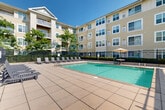 Located in Chelsea, MA, this outdoor amenity space is a standout feature of a multi-story residential complex. The centerpiece is a generously sized swimming pool with crystal-clear water, framed by a wide, tiled sundeck lined with lounge chairs — ideal for relaxing on sunny days. A black wrought-iron security fence encloses the pool area, providing safety and definition to the space.

Adjacent to the pool, a pergola-covered seating area offers shaded outdoor dining with tables and chairs, creating a comfortable retreat for residents. Umbrella-shaded tables are also positioned poolside for additional shade and convenience.

The complex features a separate landscaped courtyard, characterized by mature shade trees, mulched planting beds, and paved walkways that connect various areas of the property. Outdoor dining furniture is tucked beneath the trees, offering a quieter, more intimate gathering spot.

The surrounding multi-story building presents a classic New England aesthetic with yellow lap siding, white-trimmed multi-pane windows, and private balconies on upper floors. The thoughtful integration of green space, water features, and social areas creates a well-rounded outdoor environment that encourages community interaction and relaxation throughout the warmer months.