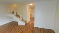 This two-story space features warm hardwood-style flooring that extends throughout the main level, creating a cohesive and inviting atmosphere. The entryway opens into a spacious layout, where a staircase with a decorative white railing serves as a notable architectural element, adding a classic touch to the interior. The stairs are carpeted, offering a pleasing contrast to the hard flooring below.

The main level flows naturally toward the kitchen area, which is visible in the background and features light wood cabinetry and neutral countertops. The kitchen is compact yet functional, with ample cabinet storage and a clean, practical design. The white walls and flush-mount ceiling light contribute to a bright, well-lit environment throughout the main floor.

The open layout between the entryway and the kitchen allows for easy movement and a sense of continuity across the space. The neutral color palette of white walls paired with warm flooring tones creates a clean, welcoming ambiance. The second floor, accessible via the staircase, adds additional living space to the overall footprint of the home.