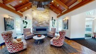 This single-story clubhouse facility is designed with a refined yet welcoming aesthetic, blending natural materials with contemporary furnishings throughout. The grand entrance greets visitors with striking herringbone hardwood flooring, soaring vaulted ceilings, and solid wood double doors flanked by sidelights that flood the foyer with natural light. A reception desk bearing the "Novo" branding anchors the entry area, setting an organized and professional tone.

The centerpiece of the space is a dramatic lounge area featuring a floor-to-ceiling stacked stone fireplace beneath an impressive vaulted ceiling with exposed wood beams and warm cedar planking. Wingback chairs and bold patterned accent seating surround a round white coffee table, creating an inviting gathering space. Large windows on either side of the fireplace provide views of the surrounding outdoor pool area and landscaping.

Adjacent to the lounge is a fully equipped kitchen and bar area, complete with granite countertops, a cooktop island with bar seating, dark cabinetry with glass uppers, a full-size stainless steel refrigerator, and a tile backsplash.

A secondary lounge offers built-in window seats, a wall-mounted TV, and additional seating. A dedicated conference room provides a long meeting table, ample seating, and large windows with outdoor views. The fitness center is well-equipped with cardio machines, free weights, floor-to-ceiling mirrors, and motivational wall graphics, all beneath wood-trimmed ceilings with natural light.