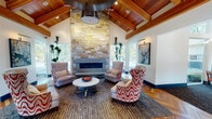 This single-story clubhouse facility is designed with a refined yet welcoming aesthetic, blending natural materials with contemporary furnishings throughout. The grand entrance greets visitors with striking herringbone hardwood flooring, soaring vaulted ceilings, and solid wood double doors flanked by sidelights that flood the foyer with natural light. A reception desk bearing the "Novo" branding anchors the entry area, setting an organized and professional tone.

The centerpiece of the space is a dramatic lounge area featuring a floor-to-ceiling stacked stone fireplace beneath an impressive vaulted ceiling with exposed wood beams and warm cedar planking. Wingback chairs and bold patterned accent seating surround a round white coffee table, creating an inviting gathering space. Large windows on either side of the fireplace provide views of the surrounding outdoor pool area and landscaping.

Adjacent to the lounge is a fully equipped kitchen and bar area, complete with granite countertops, a cooktop island with bar seating, dark cabinetry with glass uppers, a full-size stainless steel refrigerator, and a tile backsplash.

A secondary lounge offers built-in window seats, a wall-mounted TV, and additional seating. A dedicated conference room provides a long meeting table, ample seating, and large windows with outdoor views. The fitness center is well-equipped with cardio machines, free weights, floor-to-ceiling mirrors, and motivational wall graphics, all beneath wood-trimmed ceilings with natural light.