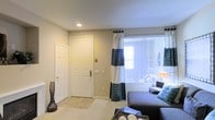 This single-floor residential space offers a well-appointed and comfortable living environment designed with both functionality and style in mind. The living area serves as the heart of the home, featuring plush carpeting in warm neutral tones that create an inviting atmosphere. A large window dressed with elegant blue and white patterned curtains and horizontal blinds allows natural light to pour into the room, lending it an airy, open feel. The space is tastefully furnished with a dark upholstered sofa accented by blue decorative pillows, complemented by a wicker-style coffee table that adds a natural texture to the room. A sleek decorative vase near the entryway adds a subtle artistic touch. The front entry is framed by a solid white paneled door, providing a clean and classic look. Recessed ceiling lighting adds a modern element while maintaining the room's warm ambiance. The neutral beige walls throughout contribute to a calm, cohesive aesthetic that makes the space feel both spacious and comfortable. Overall, this residence balances understated elegance with practical living, making it a welcoming and functional single-floor home.