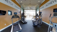This fitness center is designed to motivate and energize its users, combining functional equipment with an inviting, open atmosphere. The space is lined with rubber flooring suitable for high-traffic workout activity, and features a curated selection of cardio equipment including treadmills, an elliptical machine, and additional strength and conditioning tools such as a medicine ball station. Each cardio machine is equipped with an individual monitor, allowing users to stay entertained during their workout sessions. The walls are painted in a bold combination of orange and green, accented with large motivational words — "Challenge" and "Inspire" — that set an energetic tone throughout. Recessed ceiling lights provide bright, even illumination, while a ceiling fan helps maintain comfortable airflow. A standout architectural feature is the full-width garage-style glass door at the far end of the room, which opens directly to an outdoor patio and courtyard area, flooding the space with natural light and offering refreshing views of the surrounding exterior. This seamless indoor-outdoor connection enhances the overall ambiance, making the fitness center feel expansive and welcoming for daily use.