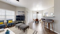 This well-designed single-floor residence offers a thoughtfully laid-out open-concept living space that seamlessly connects the living room, dining area, and kitchen. The living room is a standout feature, showcasing vaulted ceilings, wood-look flooring, and abundant natural light streaming through large windows with blinds. A ceiling fan adds comfort, and the space is styled with a navy blue leather sofa, coordinating accent chairs, and a modern multi-arm floor lamp, creating a contemporary yet inviting atmosphere.

The dining area is bathed in natural light from corner windows and features a round table with leather-upholstered chairs beneath a sleek pendant light. The kitchen is equipped with stainless steel appliances, including a refrigerator, range, microwave, and dishwasher, complemented by a classic subway tile backsplash and two-tone cabinetry. A breakfast bar with stylish bar stools provides additional seating and connects the kitchen to the main living area.

The bedrooms offer plush carpeting and ample closet space, with one room featuring an elegant four-poster bed. The bathroom includes a wide vanity with a vessel sink, gray cabinetry, a large mirror, and a shower-tub combination, all finished with wood-look flooring for a cohesive, modern aesthetic throughout.