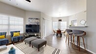 This well-designed single-floor residence offers a thoughtfully laid-out open-concept living space that seamlessly connects the living room, dining area, and kitchen. The living room is a standout feature, showcasing vaulted ceilings, wood-look flooring, and abundant natural light streaming through large windows with blinds. A ceiling fan adds comfort, and the space is styled with a navy blue leather sofa, coordinating accent chairs, and a modern multi-arm floor lamp, creating a contemporary yet inviting atmosphere.

The dining area is bathed in natural light from corner windows and features a round table with leather-upholstered chairs beneath a sleek pendant light. The kitchen is equipped with stainless steel appliances, including a refrigerator, range, microwave, and dishwasher, complemented by a classic subway tile backsplash and two-tone cabinetry. A breakfast bar with stylish bar stools provides additional seating and connects the kitchen to the main living area.

The bedrooms offer plush carpeting and ample closet space, with one room featuring an elegant four-poster bed. The bathroom includes a wide vanity with a vessel sink, gray cabinetry, a large mirror, and a shower-tub combination, all finished with wood-look flooring for a cohesive, modern aesthetic throughout.