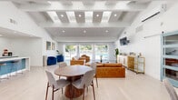 This well-appointed clubhouse in Escondido, California, offers a thoughtfully designed, open-concept layout across a single floor. The space seamlessly integrates a living room, dining area, and a fully equipped kitchen, creating an inviting environment suited for community gatherings and events.

The living room is a standout feature, boasting soaring vaulted ceilings with exposed beams and recessed lighting that enhance the sense of openness. Warm cognac leather sofas are arranged around a wood coffee table, anchored by a neutral area rug. Large sliding glass doors flood the room with natural light and provide direct views of the outdoor pool area, while a wall-mounted TV and a sleek gold bar cart add functional elegance.

The kitchen is modern and well-equipped, showcasing two-toned cabinetry in white and teal, a spacious white quartz island with bar seating for three, stainless steel appliances including a double wall oven and French door refrigerator, and a striking stainless steel range hood. White subway tile backsplash completes the clean, contemporary aesthetic.

The dining area features teal accent walls that provide bold contrast to the light wood flooring, with multiple round wooden dining tables and modern upholstered chairs. French doors and generous windows invite natural light and offer views of the surrounding greenery, creating a bright and welcoming atmosphere throughout.