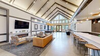 Located at 6715 Lake Shore Dr in Westmont, Illinois, this single-floor community clubhouse offers a generously sized, open-concept gathering space designed with a sleek, contemporary aesthetic. The most striking architectural feature is the dramatic vaulted ceiling with exposed beams that soar overhead, flooding the interior with natural light through expansive floor-to-ceiling windows and glass entry doors that frame views of the surrounding greenery.

The polished concrete flooring runs seamlessly throughout, tying together several distinct functional zones. The main lounge area is anchored by a cozy fireplace framed in white brick, a wall-mounted flat-screen television, and a curated seating arrangement featuring a tan leather sofa, mid-century style armchairs, and a patterned area rug. Adjacent to the lounge, a long white bar counter with wood-and-metal barstools provides a casual dining and social hub, complemented by pendant lighting above.

A dedicated dining area accommodates a large wood table with upholstered chairs beneath a striking layered pendant chandelier, ideal for group gatherings or events. A separate game and lounge nook features a shuffleboard table and additional seating surrounded by bold, framed abstract artwork. Dark horizontal banding on the walls throughout adds graphic contrast and visual cohesion to the bright, airy interior, creating a polished and inviting community environment.