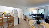 This single-floor, three-bedroom, one-bathroom unit features an open-concept layout that efficiently connects the main living areas. The kitchen is positioned adjacent to the living and dining spaces, separated by a half-wall counter that doubles as a breakfast bar with seating. White cabinetry and a white refrigerator give the kitchen a clean, bright appearance, with the word "kitchen" displayed as a charming decorative accent above the entryway.

The living area features neutral-toned carpeting and is furnished with a sectional sofa, coffee table, and additional seating, creating a comfortable gathering space. A window along the far wall allows natural light to filter into the room, brightening the overall atmosphere. The dining area sits just off the living space and accommodates a round wooden table with chairs, making it suitable for everyday meals.

The layout flows naturally from one area to the next, maximizing the use of the available footprint. Crown molding adds a subtle architectural detail throughout the shared spaces. Overall, the unit offers a practical and livable environment ideal for those seeking a cozy, well-connected floor plan.
