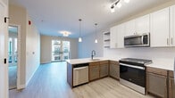 Located at 101 S Main St in Lombard, Illinois, this well-appointed single-floor residence offers a thoughtfully designed open-concept layout that seamlessly connects the kitchen and living area. The kitchen is a standout feature, showcasing a two-tone cabinet design with white upper cabinetry and warm wood-toned lower cabinets, all accented with gold hardware. A peninsula with a white countertop and undermount sink provides additional workspace and casual seating potential, while stainless steel appliances — including a gas range, built-in microwave, and dishwasher — complete the cook's setup. Pendant lighting above the peninsula and track lighting overhead add layers of illumination. Light wood-style flooring flows throughout the main living areas, contributing to a bright, airy atmosphere. The living area extends toward a set of glass doors that open to a private balcony, allowing natural light to pour in and offering views of the surrounding urban streetscape. The bedroom features plush carpeting and a large window that further enhances natural light. The bathroom is finished with crisp white cabinetry, a dark countertop, matte black fixtures, and a soaking tub with an integrated shower. A smart thermostat adds a modern, connected touch to this contemporary Lombard residence.