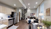 This two-story space combines modern design with functional living across an open, well-connected floor plan. The main level integrates a kitchen and living area in a seamless open-concept layout. The kitchen is equipped with stainless steel appliances, including a full-size refrigerator and dishwasher, complemented by light wood cabinetry and a classic stove. Dark hardwood-style flooring runs throughout, adding warmth and continuity to the space.

A standout architectural feature is the elegant wrought-iron spiral staircase, which serves as both a functional element and a visual centerpiece, connecting the main level to the upper floor with a sculptural presence. Recessed lighting is installed throughout the ceiling, providing consistent, modern illumination across the open space.

The living area is generously proportioned, accommodating a full seating arrangement including a white sofa and accent chairs, alongside a media console and decorative artwork. Natural light filters in through a window on the far wall. The overall ambiance is contemporary and polished, with neutral gray walls and dark flooring creating a cohesive, sophisticated aesthetic throughout.