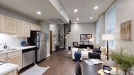 This two-story space combines modern design with functional living across an open, well-connected floor plan. The main level integrates a kitchen and living area in a seamless open-concept layout. The kitchen is equipped with stainless steel appliances, including a full-size refrigerator and dishwasher, complemented by light wood cabinetry and a classic stove. Dark hardwood-style flooring runs throughout, adding warmth and continuity to the space.

A standout architectural feature is the elegant wrought-iron spiral staircase, which serves as both a functional element and a visual centerpiece, connecting the main level to the upper floor with a sculptural presence. Recessed lighting is installed throughout the ceiling, providing consistent, modern illumination across the open space.

The living area is generously proportioned, accommodating a full seating arrangement including a white sofa and accent chairs, alongside a media console and decorative artwork. Natural light filters in through a window on the far wall. The overall ambiance is contemporary and polished, with neutral gray walls and dark flooring creating a cohesive, sophisticated aesthetic throughout.