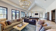Located at 5252 Riggins Run in Dublin, Ohio, this expansive single-floor community clubhouse offers a generously proportioned gathering space designed with upscale finishes and thoughtful architectural detail. The entry foyer immediately impresses with its dramatic vaulted ceiling accented by exposed wood arch beams, a striking herringbone-patterned hardwood floor, and a crystal chandelier suspended at its center. Flanking the entryway are built-in cabinetry stations and a dedicated workstation area, providing functional amenities alongside refined aesthetics. Stacked stone accent columns frame the corridor leading to the main living area, where floor-to-ceiling glass doors invite abundant natural light and outdoor views.

The main lounge area is anchored by a towering stacked-stone fireplace with a wood mantle and linear firebox, complemented by soaring cathedral ceilings, a statement candelabra chandelier, and multiple seating arrangements suited for both relaxed conversation and social gatherings. A billiards table and wall-mounted flat-screen televisions add entertainment options throughout the space. Dark-framed windows and doors provide a bold contrast to the bright white walls, while oversized windows offer views of the surrounding community and pool area. A substantial wooden bookcase cabinet adds a classic, library-inspired touch. The overall ambiance blends modern elegance with warm, transitional design elements.