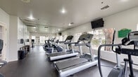 This single-level fitness center provides a dedicated workout space designed to accommodate a variety of cardio exercises. The room features durable rubber flooring throughout, providing a practical and resilient surface suited for exercise equipment. Large multi-pane windows line the walls, flooding the space with natural light and offering pleasant views of the surrounding outdoor greenery, creating an open and motivating atmosphere.

The facility is equipped with multiple treadmills, an elliptical trainer, a stationary bike, and additional cardio machines, all arranged to maximize the available floor space. Full-length mirrors along one wall visually expand the room and allow users to monitor their form during workouts. A wall-mounted flat-screen television is positioned for easy viewing from the cardio equipment.

Recessed and flush-mounted ceiling lights supplement the natural light, ensuring the space remains well-lit throughout the day. A wall-mounted rack and storage accessories are available near the entry for equipment organization. Access to the fitness center is provided through a glass-paned French door, which connects to an adjacent common area. Ventilation is supported by ceiling-mounted HVAC vents, maintaining a comfortable environment during exercise.