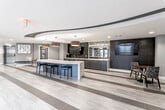 This contemporary amenity space is designed with a polished, upscale aesthetic that balances functionality with sophisticated style. The open layout is anchored by a striking curved ceiling element with an integrated dark trim detail, which adds architectural depth and visual interest to the main lounge area. Large-format light wood-toned tile flooring flows seamlessly throughout, unifying the various zones of the space.

At the center of the lounge, a substantial island with a marble-veined white stone countertop serves as a focal point, surrounded by sleek black bar stools. Adjacent to it, a full wet bar runs along one wall, finished with matching stone counters, dark cabinetry, an undermount sink, and a wall-mounted television set against a dramatic textured black tile accent wall with an antiqued mirror backdrop.

A seating nook provides an intimate gathering area, complete with a round wood table, patterned chairs, a wall-mounted display screen, and vibrant abstract artwork. Pendant lighting with warm gold interiors adds a rich contrast to the cool-toned palette throughout.

Near the entrance, a polished stainless steel elevator, a tufted black-and-gold bench, and curated wall art create a refined welcome. Floor-to-ceiling windows offer views of an outdoor courtyard and pool area, bringing in natural light and reinforcing the connection to exterior amenities.