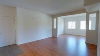 This single-floor space offers a straightforward and functional layout with a welcoming, light-filled atmosphere. The interior is finished with warm hardwood-style flooring that runs throughout, lending a sense of continuity and cohesion across the connected living areas. Crisp white walls reflect natural light generously, creating an airy and open ambiance throughout the space. The main living area flows seamlessly into an adjacent room through a wide open archway, making the overall floor plan feel spacious and interconnected. A window in the adjoining room allows natural light to stream in, brightening the interior and providing a pleasant view to the outside. The clean, neutral palette of the walls and flooring provides a versatile backdrop well-suited for a variety of furnishing styles and personal touches. The smooth ceilings and minimal architectural detailing give the space a modern, uncluttered feel. Overall, this single-level space is characterized by its open connectivity, consistent finishes, and bright, welcoming atmosphere.