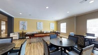 This single-floor amenity space is thoughtfully designed to serve as a social hub for recreation and relaxation. The open layout accommodates both casual lounging and active entertainment, creating a versatile environment suited to a variety of gatherings.

The room features warm yellow accent walls complemented by neutral taupe tones, crowned with classic crown molding that adds a refined architectural touch. Recessed lighting in the white ceiling provides a bright, even illumination throughout the space. Natural light flows in through a window dressed with white shutters, enhancing the welcoming ambiance.

A centerpiece of the room is a rich mahogany shuffleboard table, which serves as a focal point for recreational activity. Surrounding it are multiple round and rectangular dining tables paired with upholstered blue-gray chairs, offering comfortable seating for guests. A cushioned bench lines one wall, adding additional lounge seating options.

Light wood-toned laminate flooring runs throughout, grounding the space with a warm, contemporary feel. Decorative abstract artwork in soft, muted tones adorns the walls, contributing to the polished yet approachable aesthetic. The overall design balances functionality with style, making this an inviting space for socializing, gaming, and community engagement.