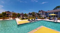 This amenity space combines outdoor resort-style living with a well-appointed indoor fitness facility. The centerpiece is a sparkling outdoor pool featuring bright yellow accent elements, in-pool lounge chairs for sunbathing at water level, and a generous surrounding deck furnished with additional chaise lounges and seating areas. A covered pergola structure anchors a landscaped lounge zone, enclosed by black aluminum fencing, with manicured green space and mature trees completing the open-air setting beneath clear blue skies.

The fitness center is divided into distinct functional zones. A dedicated cardio room features multiple treadmills positioned beneath wall-mounted televisions, with large windows on multiple walls flooding the space with natural light and views of the surrounding community. The main strength training area is a generously sized, open room finished with patterned herringbone-style carpet and lined with full-length mirrors that amplify the sense of space. Equipment includes a comprehensive cable and power rack system, a full dumbbell rack, medicine ball storage, battle ropes, stability balls, a rowing machine, a recumbent cycle, and additional cable machines. Ceiling fans and recessed lighting maintain a comfortable environment throughout, while windows provide natural light and outdoor visibility on multiple sides.