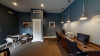 Located at 9611 Grant Rd in Houston, Texas, this single-floor office space combines professional functionality with a welcoming, well-appointed interior design. The space is unified by rich teal-blue walls throughout, complemented by warm carpeting in earthy tones that add depth and comfort underfoot.

The workspace area features a substantial dark wood workstation along one wall, equipped with multiple computer monitors and office equipment, illuminated by a pair of stylish industrial-style pendant lights that hang overhead. Ergonomic executive chairs and additional seating with a small table provide space for collaborative work or client meetings.

A comfortable lounge area occupies one corner of the space, furnished with a deep burgundy sofa accented by zebra-print throw pillows and a pair of classically striped armchairs. A large window with layered blinds and striped sheer curtains allows natural light to filter in while offering views of the surrounding greenery outside.

The interior decor carries a distinct Texas character, highlighted by a decorative wooden Texas state cutout mounted on the wall, alongside framed Western artwork and colorful city-themed prints. Recessed ceiling lighting and pendant fixtures ensure the space remains well-lit throughout. A louvered interior door provides access to an additional room, rounding out this thoughtfully designed professional environment.