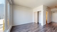 This single-floor unit presents a clean, modern aesthetic throughout, unified by durable grey-toned hardwood-style flooring that extends across all living areas. The contemporary kitchen is efficiently designed, featuring white cabinetry paired with a warm natural wood open shelf, grey subway tile backsplash, a deep farmhouse-style sink, and a full suite of stainless steel appliances, including a refrigerator, dishwasher, microwave, and stove. The bedroom offers a generously sized, open layout with a built-in sliding-door closet providing ample storage. A wall-mounted heating unit ensures year-round comfort. A large picture window in the living area fills the space with natural light while offering views of the surrounding neighborhood. The bathroom is finished with a clean, white tub and shower combination featuring chrome fixtures, maintaining the unit's crisp, minimal design language. The overall layout, as seen in the floorplan overview, is compact yet functional, with clearly defined spaces and straightforward flow between rooms. Bright white walls throughout contribute to an open, airy atmosphere that complements the modern finishes.