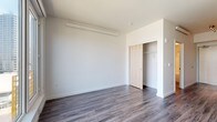 This single-floor unit presents a clean, modern aesthetic throughout, unified by durable grey-toned hardwood-style flooring that extends across all living areas. The contemporary kitchen is efficiently designed, featuring white cabinetry paired with a warm natural wood open shelf, grey subway tile backsplash, a deep farmhouse-style sink, and a full suite of stainless steel appliances, including a refrigerator, dishwasher, microwave, and stove. The bedroom offers a generously sized, open layout with a built-in sliding-door closet providing ample storage. A wall-mounted heating unit ensures year-round comfort. A large picture window in the living area fills the space with natural light while offering views of the surrounding neighborhood. The bathroom is finished with a clean, white tub and shower combination featuring chrome fixtures, maintaining the unit's crisp, minimal design language. The overall layout, as seen in the floorplan overview, is compact yet functional, with clearly defined spaces and straightforward flow between rooms. Bright white walls throughout contribute to an open, airy atmosphere that complements the modern finishes.
