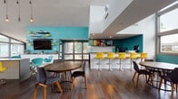 This dynamic single-floor clubhouse is designed with a bold, contemporary aesthetic that immediately captures attention. Signature teal walls, vivid yellow accents, and dark hardwood flooring run throughout, creating a cohesive and energetic atmosphere.

The main lounge area features soaring ceilings with clerestory windows that flood the space with natural light. Eclectic furnishings—including plush lounge chairs, a sectional sofa, and a geometric-patterned area rug—are complemented by striking wall art, including a notable colorful skull canvas. A bold graphic-print accent wall frames a teal standing desk with yellow barstools, providing a dedicated workstation zone.

Adjacent to the lounge, a stylish bar counter with white tile detailing is lined with bright yellow pedestal barstools and features colorful artwork as a backdrop. A comfortable dining area with a round wood table offers additional seating nearby.

A dedicated game room provides a round poker-style table surrounded by upholstered chairs, with large windows overlooking lush tree canopies. The main hall opens to a full-size billiards table beneath a cluster of Edison-style pendant lights, set against a bold striped feature wall in teal, yellow, and gray.

The kitchen area is equipped with a full-size refrigerator, microwave, and ample cabinetry, supporting the space's function as a fully amenitized gathering destination.