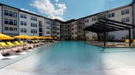 This amenity space seamlessly blends sophisticated indoor and outdoor living into a cohesive, resort-inspired environment. At its heart is a generously sized swimming pool with crystal-clear blue water, set within a courtyard framed by a multi-story building on multiple sides. The pool deck is well-appointed with rows of white lounge chairs, vibrant yellow umbrellas for shade, and a sleek dark-framed pergola that adds architectural interest and additional covered seating.

Adjacent to the pool, a covered outdoor lounge area offers a comfortable transition between inside and out, featuring stylish striped area rugs, plush seating, and direct views of the pool through wrought iron fencing. Stamped concrete paving adds texture and visual appeal to the terrace.

The interior lobby is a standout, boasting soaring double-height ceilings with clerestory windows that flood the space with natural light. Polished marble-look flooring reflects the light beautifully, while rich green velvet sofas, wood accent tables, and white cylindrical ottomans create an inviting seating arrangement. A sleek fireplace anchors one wall, and curated wall art adds character. Lush fiddle-leaf fig trees in modern grey planters provide organic warmth near the entryway, connecting the refined interior to the vibrant outdoor amenity beyond.