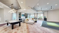 Located at Integra TC in Daytona Beach, FL, this single-floor community clubhouse offers a generously proportioned, open-concept layout designed for socializing and recreation. The main living area features soaring vaulted and tray ceilings, recessed lighting, and a ceiling-mounted projector, creating a versatile entertainment hub. A large sectional sofa anchors the lounge space, while oversized windows dressed in elegant drapes flood the room with natural light and offer views of the Florida landscape.

The kitchen and dining zone are anchored by a lengthy white quartz island with bar seating, complemented by eye-catching globe and halo pendant chandeliers in gold finishes. Built-in open shelving with gold accents, a mounted flat-screen TV, and a wet bar with a deep basin sink add functionality and flair. Wood-look tile flooring grounds the kitchen area, while the accent wall in deep burgundy adds a bold design statement.

A dedicated game room provides air hockey, billiards, and foosball tables beneath a wood-paneled tray ceiling with contemporary globe chandeliers. High-top bistro tables positioned near floor-to-ceiling windows offer casual seating throughout. A standout indoor golf simulator room, complete with a projection screen and putting turf, rounds out the recreational amenities, making this a truly dynamic and well-appointed community gathering space.