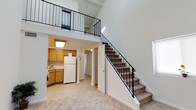 This two-story residence is defined by its dramatic vertical design, with soaring ceilings that create an expansive, airy atmosphere throughout the main level. The open layout connects the living area to the kitchen seamlessly, allowing for an easy flow between spaces.

The kitchen is fitted with warm honey-toned wood cabinetry offering generous storage, a white refrigerator, and light-toned flooring. A flush-mounted ceiling light provides functional illumination. An adjacent doorway leads further into the unit, suggesting additional rooms beyond the main living space.

The main living area features neutral carpet flooring, contributing to a soft and comfortable underfoot feel. A carpeted staircase with a sleek black metal railing ascends to the upper level, which features an open loft-style layout overlooking the main floor below. The loft is enclosed by matching black metal railings, maintaining a cohesive design aesthetic while preserving the open sightlines from below.

The combination of high ceilings, an open loft, and interconnected living spaces gives this two-story residence a notably spacious and functional character.