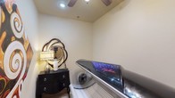 The tanning room is a compact, purpose-built space designed to provide a private and comfortable indoor tanning experience. At its center sits an Ergoline tanning bed, a sleek, modern unit with a distinctive dark metallic finish and illuminated panel, offering a professional-grade tanning session within the comfort of the space. A small portable fan is positioned nearby, ensuring adequate airflow and comfort during use.

The room's warm, neutral-toned walls create a calm and relaxing atmosphere. A ceiling fan overhead provides additional ventilation and contributes to a pleasant environment. Tasteful decorative touches elevate the space beyond purely functional use — a bold, ornate circular mirror with a layered sunburst-style frame adorns one wall, complemented by a classic black dresser beneath it, topped with a table lamp and small accessories that lend a refined, boutique-like feel.

The tile flooring is easy to maintain and practical for the room's purpose. Overall, the tanning room balances functionality with thoughtful décor, creating a welcoming, salon-quality environment suited for regular use.